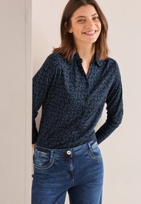 Camicia button-up blu navy con motivo astratto blu chiaro; abbinata a jeans blu scuro con cuciture decorative e tasche frontali.