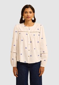 Camicia bianca con ricami floreali blu, maniche lunghe e dettagli a inserti, realizzata in un tessuto leggero.