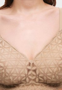 Bežový krajkový bralet se květinovým vzorem. Má výstřih do V a vlnité okraje. Hladká textura s nastavitelnými ramínky.