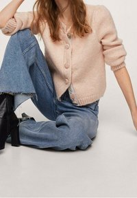 Jasnoróżowy sweter na guziki z okrągłym dekoltem, zestawiony z niebieskimi jeansami o szerokich nogawkach. Denim jest lekko postrzępiony na dole.