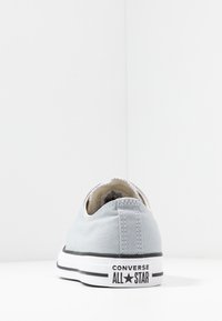 Baskets en toile grises avec semelle en caoutchouc blanche, featuring un détail de rayure noire et le logo "Converse All Star" sur le talon. Design minimaliste.