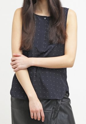 Kvinde med langt mørkt hår iført en ærmeløs marineblå top med prikker og sort nederdel, armene krydsede over kroppen.