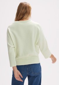 Femme aux cheveux longs portant un pull ample côtelé vert clair et un jean bleu, photographiée de dos sur un fond blanc uni.