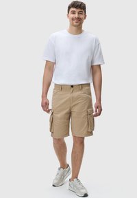 Beige cargo shorts med flere sidelommer, knappestengning og avslappet passform, kombinert med en enkel hvit t-skjorte og sportssko.