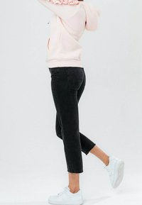 Sudadera con capucha de color rosa claro con detalles de volantes, combinada con vaqueros negros cortos. La modelo lleva zapatillas deportivas blancas. Diseño de conjunto sencillo y casual.