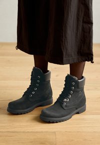 Botas de tobillo de ante negro con cuello de piel, puntera redonda, cordones en la parte delantera y suela de goma resistente, situadas sobre un suelo de madera.