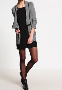 Schwarzes Kleid mit einem gestreiften, offen vorne getragenen Cardigan mit langen Ärmeln. Der Cardigan hat kontrastierende weiße Streifen und eine lockere Passform.
