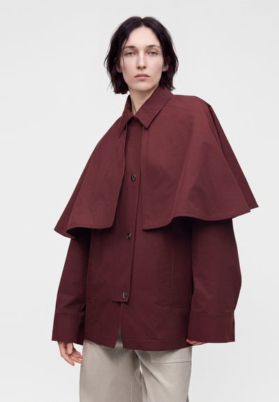 Personne aux cheveux courts et foncés portant une veste rouge foncé oversized avec un large col cape et un pantalon beige clair, debout devant un fond uni.