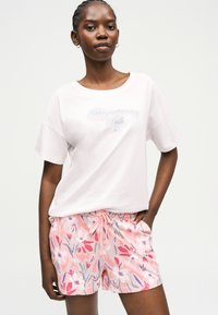 Γυναίκα που φοράει λευκό T-shirt με γαλάζια γραμματοσειρά και ροζ σορτς με λουλούδια, στέκεται με το ένα χέρι στην τσέπη μπροστά σε ένα απλό φόντο.