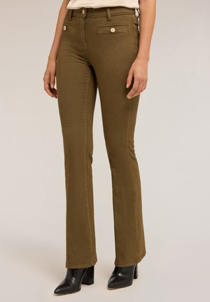 Pantalons évasés vert olive avec une texture lisse, deux poches avant et des accents métalliques sur la taille, associés à des bottes à talons noires.