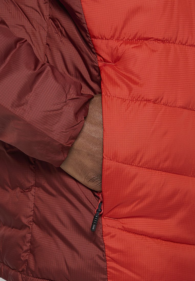 Ręka w kieszeni czerwonej i bordowej pikowanej kurtki puchowej z widocznym uchwytem suwaka.