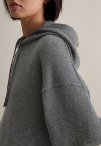 Grauer gestrickter Kapuzenpullover mit Kordelzug, der einen lockeren Schnitt, weite Ärmel und eine weiche Textur aufweist. Der Stoff wirkt warm und bequem.
