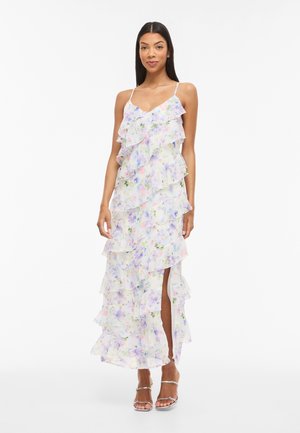 Vrouw draagt een witte maxi-jurk met bloemenprint, ruches, spaghettibandjes en een split aan de voorkant, gecombineerd met zilveren open-toe hakken.