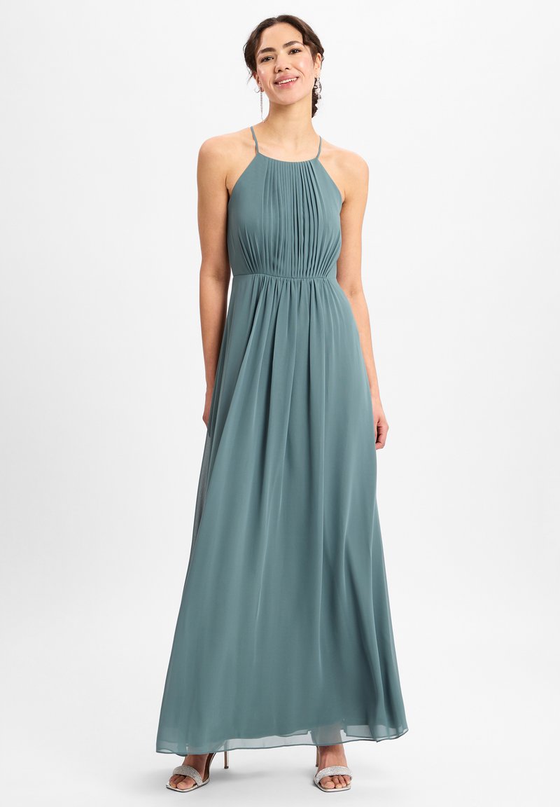 Een lange, vloeiende halterneck jurk van zacht teal stof, met verticale plooien en een nauwsluitende taille, aangevuld met delicate strapsandalen.
