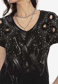 Camiseta negra con manchas plateadas y diseños de pinceladas. Cortes desgastados en las mangas y acentos de rhinestones añaden textura.