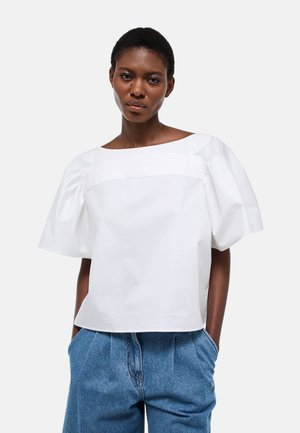 Femme portant un chemisier blanc à manches bouffantes et un pantalon en denim bleu, debout les mains dans les poches devant un fond uni.