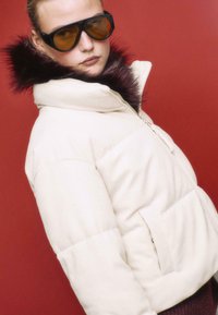 Puffermantel in Creme mit hohem Kragen und einem dunklen Kunstfellbesatz; übergroße Taschen und gerippte Textur; das Model trägt große Sonnenbrillen.