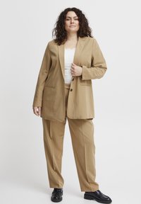 Barna csíkos öltöny, szabott blézerrel és magas derekú nadrággal, sima anyagból készült. Kiegészítők között fekete cipők találhatók.