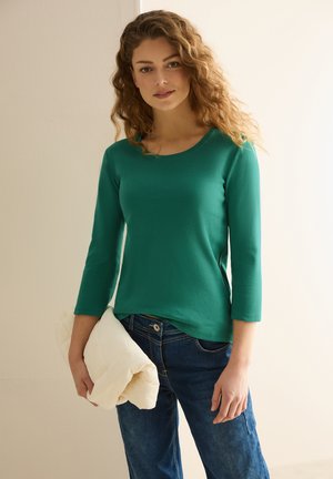Haut ajusté à manches longues de couleur verte en tissu doux, associé à un jean bleu. Le modèle tient un sac matelassé de couleur crème. Design et couleurs simples.