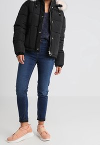 Person som bär en svart vadderad jacka med pälskant på huvan, blå jeans, lila stickad tröja och rosa slip-on-sneakers, stående mot en enfärgad bakgrund.