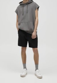 Sweat à capuche gris sans manches avec poche avant, associé à un short noir. Chaussures blanches, complétées par des chaussettes grises. Design simple et décontracté.
