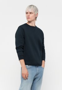 Felpa navy scuro in un tessuto morbido e liscio, con colletto a girocollo, maniche lunghe e polsini a costine. Abbinata a jeans azzurri sdruciti.