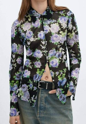 Vrouw draagt een transparante zwarte blouse met paarse bloemenprint, deels dichtgeknoopt, ingestopt in een blauwe spijkerbroek met een zwarte riem.