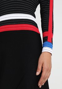 Lähikuva henkilöstä, jolla on päällään musta ribbimekko punaisilla, valkoisilla ja sinisillä raidallisilla pitkillä hihoilla sekä näkyvillä oikeilla käsillä.