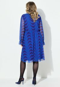 Robe bleue, longueur genou, avec une superposition en voile à motif ondulé, taille élastique et manches longues. Associée à des chaussures à talons assorties.