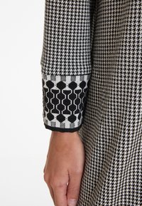 Schwarz-weißer Hahnentrittmusterstoff mit einem gemusterten Bündchen, das geometrische Motive zeigt. Glatte Textur, taillierter Schnitt, Nahaufnahme von Ärmel und Hand.