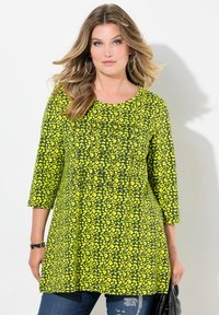 Grøn tunika med gult leopardprint, rund halsudskæring, trekvartærmer og en let udvidet pasform. Sammen med mørke jeans og en sort taske.