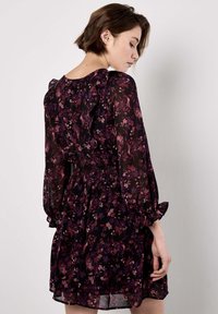 Schwarzes Blumenkleid mit lila und rosa Blütenblättern, gerafften Ärmeln, elastischem Bund und fließendem Schnitt, gefertigt aus leichtem, durchscheinendem Stoff.