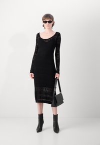 Abito nero a maglia con maniche lunghe, caratterizzato da un motivo testurizzato. Abbinato a stivaletti neri alla caviglia e a una borsa strutturata.