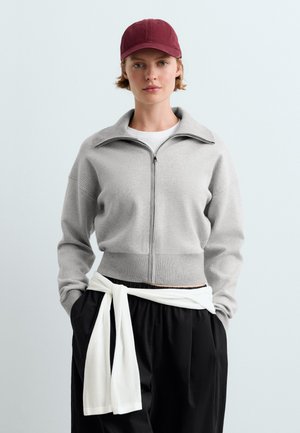 Jeune personne portant une casquette bordeaux, un pull gris zippé, une chemise blanche nouée autour d’un pantalon noir, debout les mains dans les poches devant un fond clair.