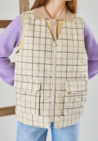 Vadderad beige väst med rutmönster, dragkedja framtill, två frontfickor och en brun krage, bärs över en lila långärmad topp.