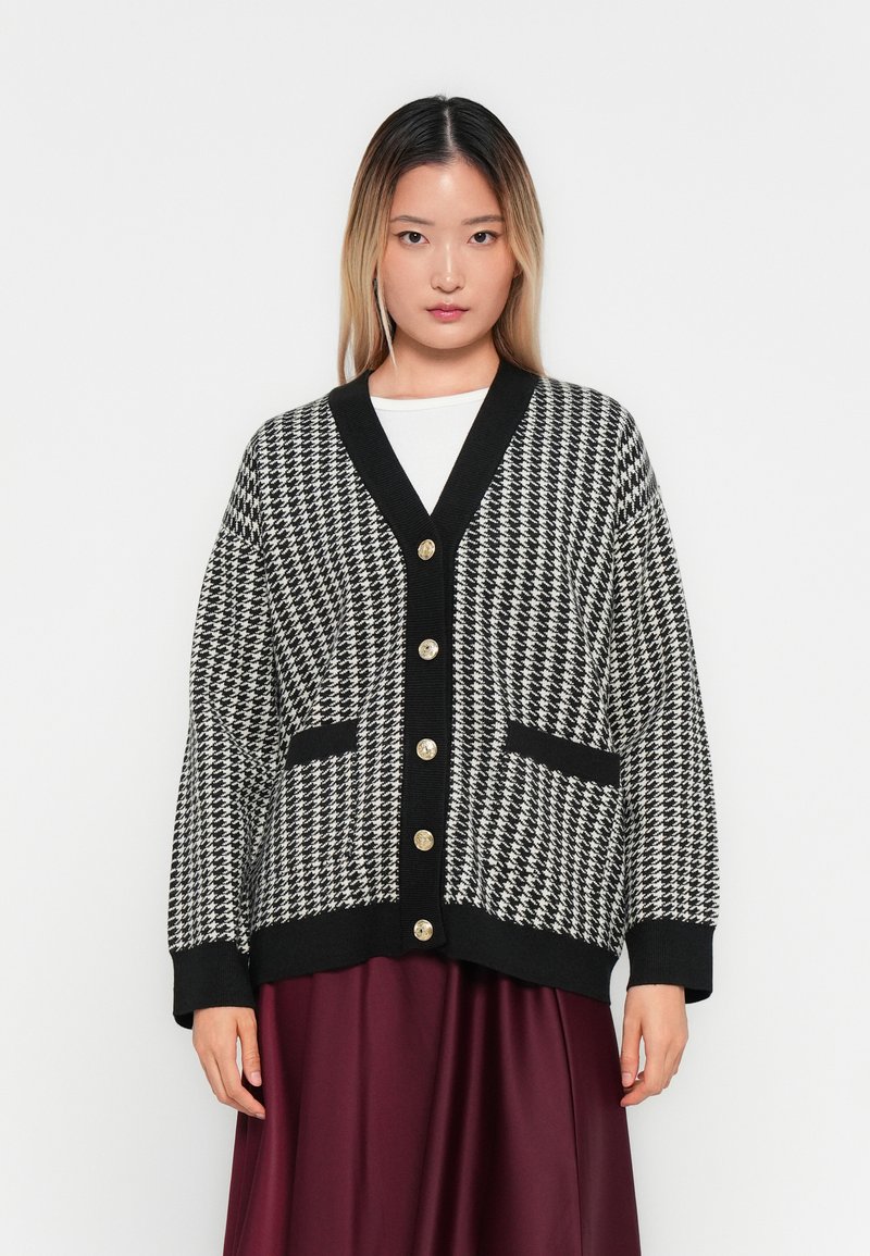 Woman wearing black and white houndstooth cardigan with gold buttons over white top and burgundy skirt, standing against a white background.