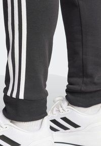 Schwarze Sweatpants mit drei weißen Streifen an den Seiten. Kombiniert mit weißen Sneakers mit schwarzen Akzenten und einem strukturierten Mesh-Obermaterial.