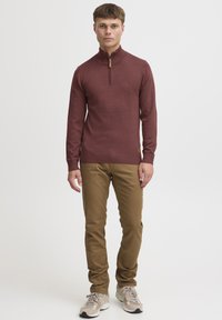 Sweat à fermeture éclair bordeaux avec un col côtelé, associé à un pantalon kaki. L'ensemble est complété par des baskets de couleur claire.
