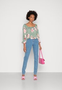 Blusa floral verde com mangas bufantes, combinada com jeans azul claro. Os acessórios incluem uma bolsa rosa e saltos rosa combinando.