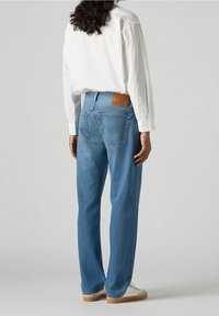 Vaqueros de mezclilla de color azul claro con un corte recto, bolsillos traseros y un parche de cuero marrón en la cinturilla. Combinados con una camisa blanca.