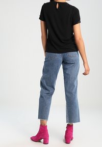 Mujer con una blusa negra de manga corta, jeans cortos azules y botas de ante rosa brillante de tobillo, de pie con la espalda hacia la cámara.