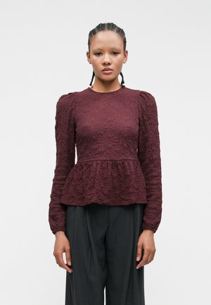 Bordo textured bluza s dugim rukavima i peplum strukom. Ima okrugli izrez i nabrané pufaste ramena. Uparena s tamnim hlačama.