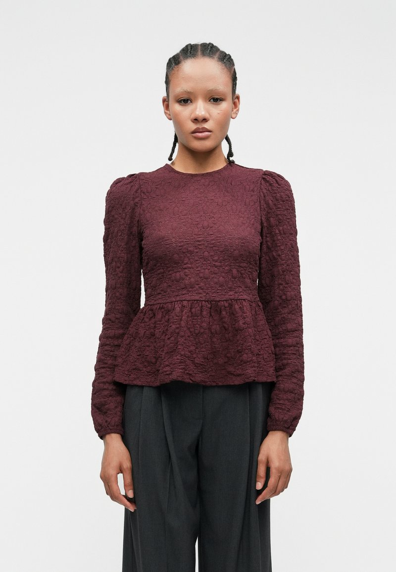 Blusa borgogna texturizzata con maniche lunghe e vita a peplum. Presenta un collo a giro e spalle a sbuffo arricciate. Abbinata a pantaloni scuri.