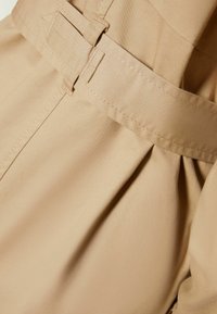 Vêtement beige avec une texture lisse, doté d'une ceinture avec une boucle rectangulaire et un détail plissé à la taille.