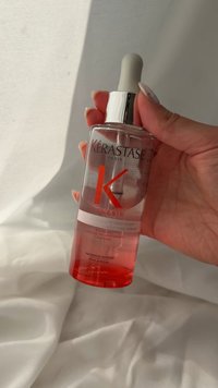 Hånd, der holder Kérastase Genesis hårstyrkende serumflaske med pipette, pink væske indeni, på hvidt stofbaggrund.