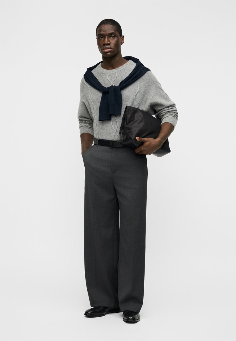 Pull gris en tricot avec motif torsadé, écharpe bleue marine nouée autour du cou, pantalon gris taille haute, ceinture noire, tenant une pochette noire.