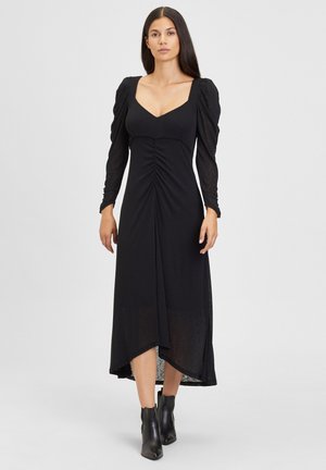 Abito nero con maniche lunghe e voluminose, scollatura a V profonda, vita cintrata e orlo asimmetrico. Realizzato in materiale leggero. Abbinato a stivaletti.