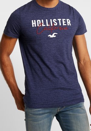 Mężczyzna ubrany w granatowy T-shirt Hollister California i jasnoniebieskie dżinsy, stojący z jedną ręką opartą na biodrze.