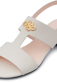 Sandal in beige chiaro con una fascia larga, design con ritagli e un accento decorativo dorato. La suola è nera con una soletta rosa per creare contrasto.