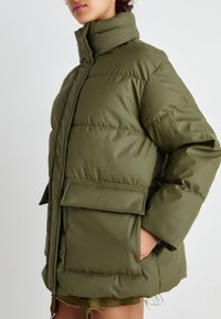 Veste rembourrée vert olive avec un col haut, fermeture éclair, grandes poches avant et un design matelassé subtil. Tissu lisse et texturé.